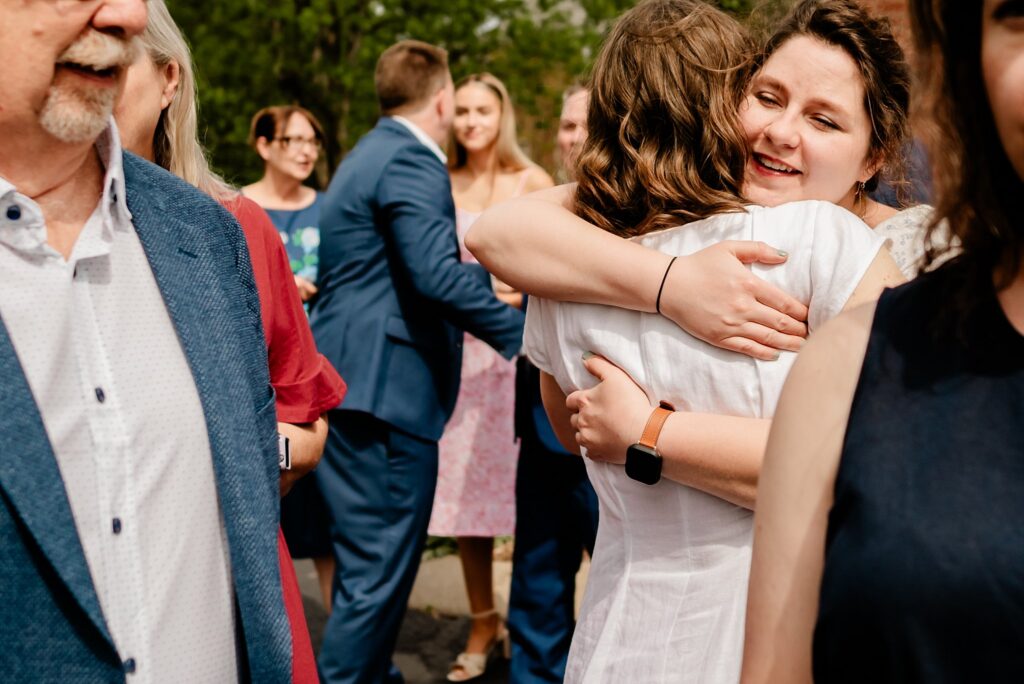A woman wearing a watch smiles while hugging another woman outdoors. Several people in formal clothing stand nearby, some talking and others embracing, suggesting a celebratory event—captured candidly by a Nashville wedding photographer amid the trees.