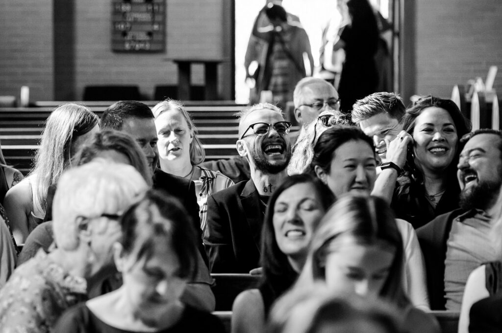 A group of people sit closely together on church pews, laughing and smiling. Captured by a Nashville wedding photographer, the joyful atmosphere is lively as men and women enjoy a special moment during the gathering or event.