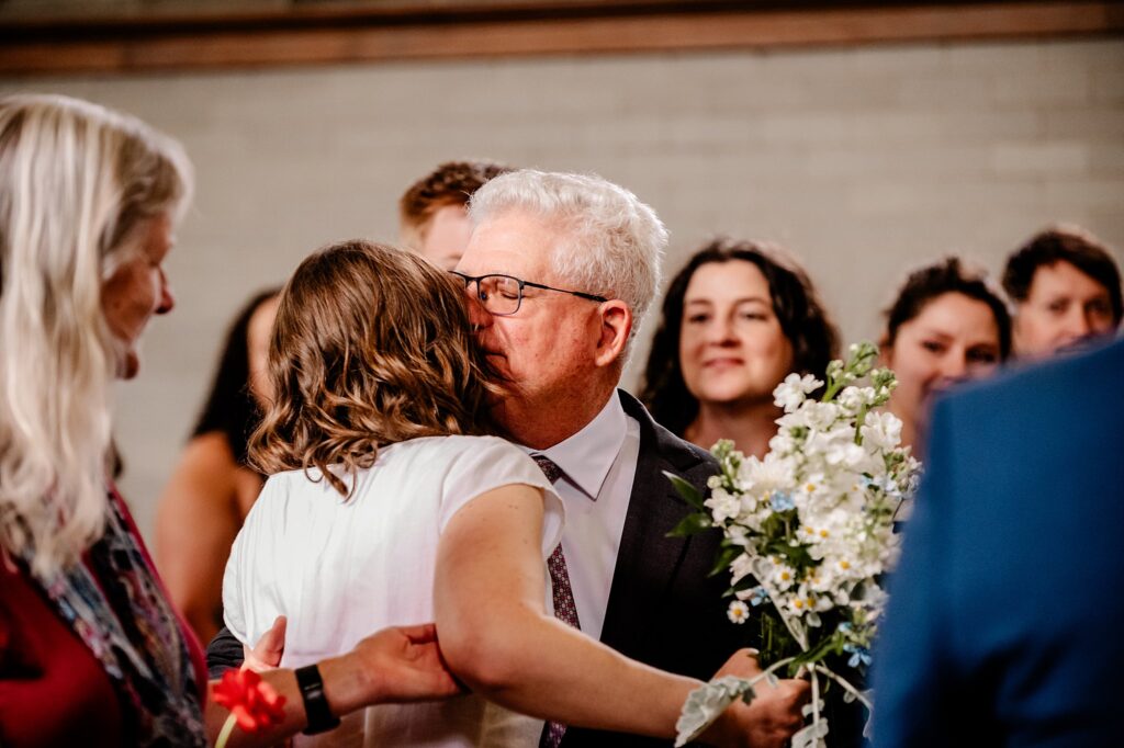 A man with gray hair and glasses hugs a woman in a white dress holding a bouquet, surrounded by smiling guests—captured beautifully by a Nashville wedding photographer at a joyful celebratory event.