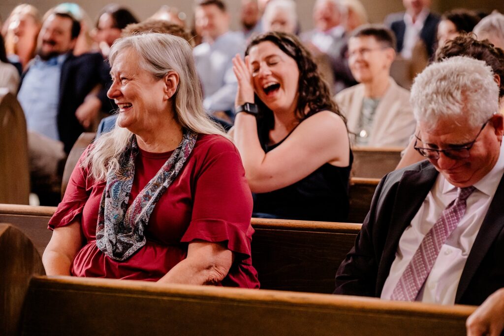 A group of people sit in wooden pews, smiling and laughing. A woman in a maroon top and scarf sits in front, while others behind her appear happy—captured candidly by a Nashville wedding photographer during an indoor event.