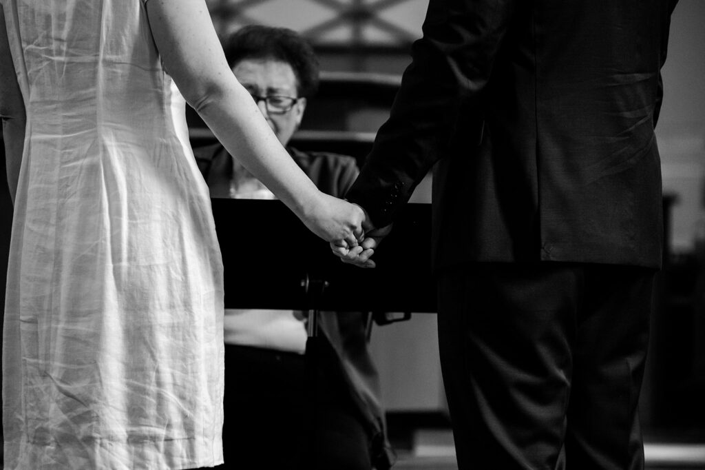 A couple holding hands, facing away from the camera, with a person sitting in the background, possibly officiating a ceremony. Captured in black and white by a Nashville wedding photographer.