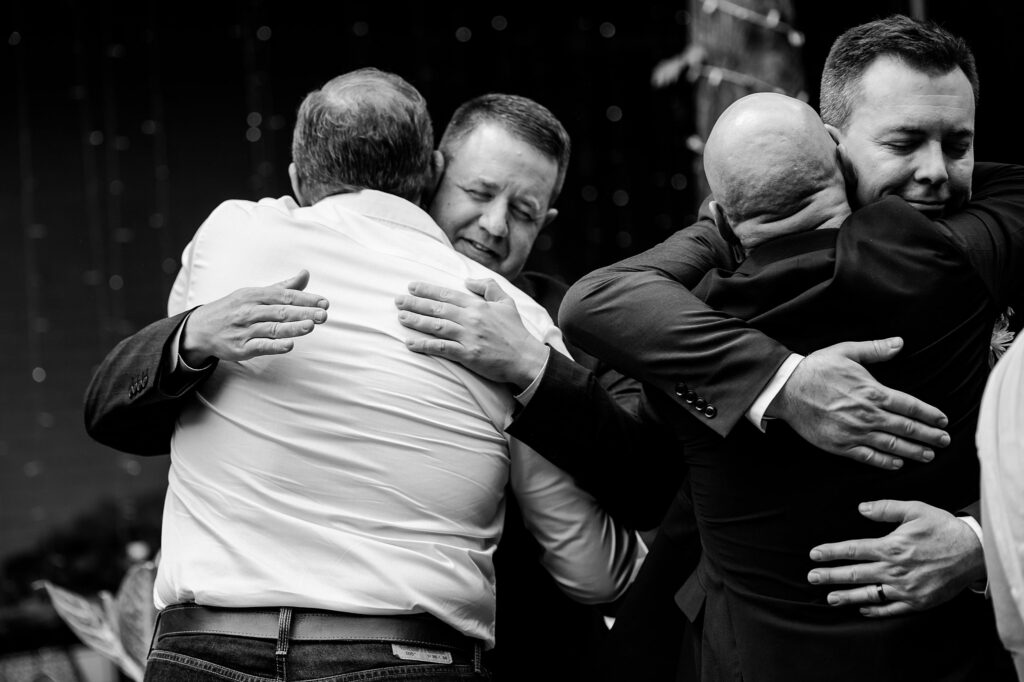 Two pairs of men in suits embrace each other in heartfelt hugs. This black-and-white scene, captured by a Nashville wedding photographer, highlights a powerful moment of connection and support.