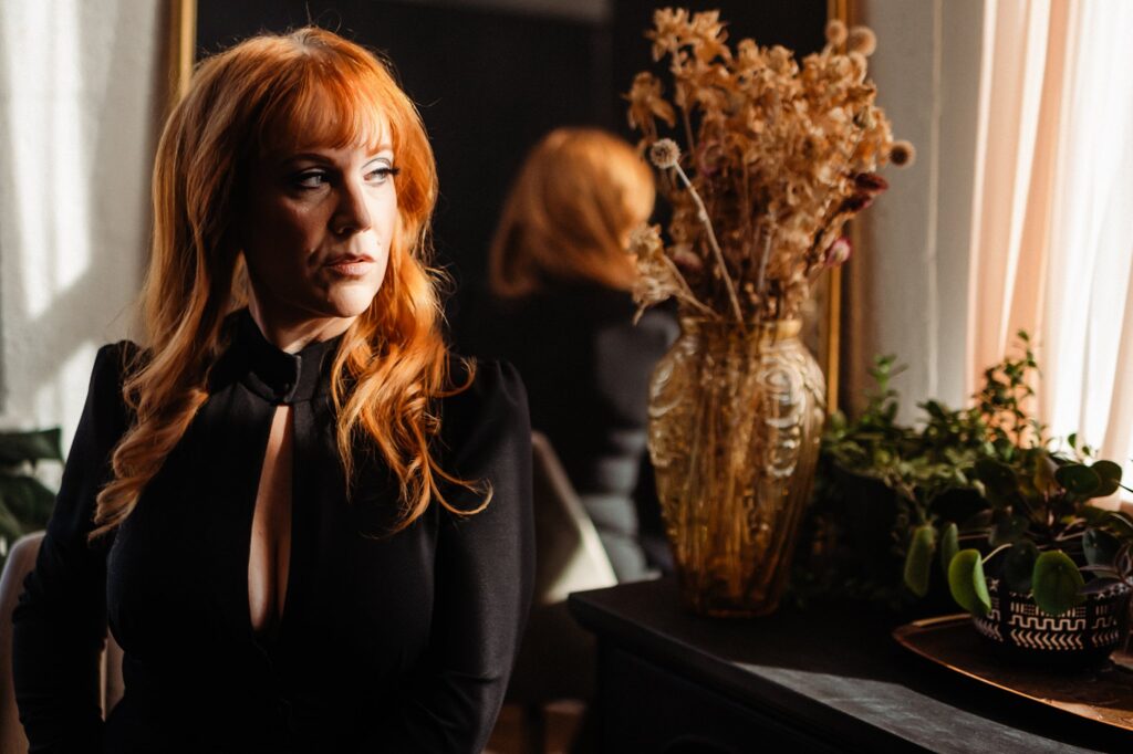A woman with long red hair and bangs, dressed in black, sits indoors near a window. Dried flowers and a green plant rest nearby, while her reflection appears in a mirror—captured by a Nashville creative portrait photographer.