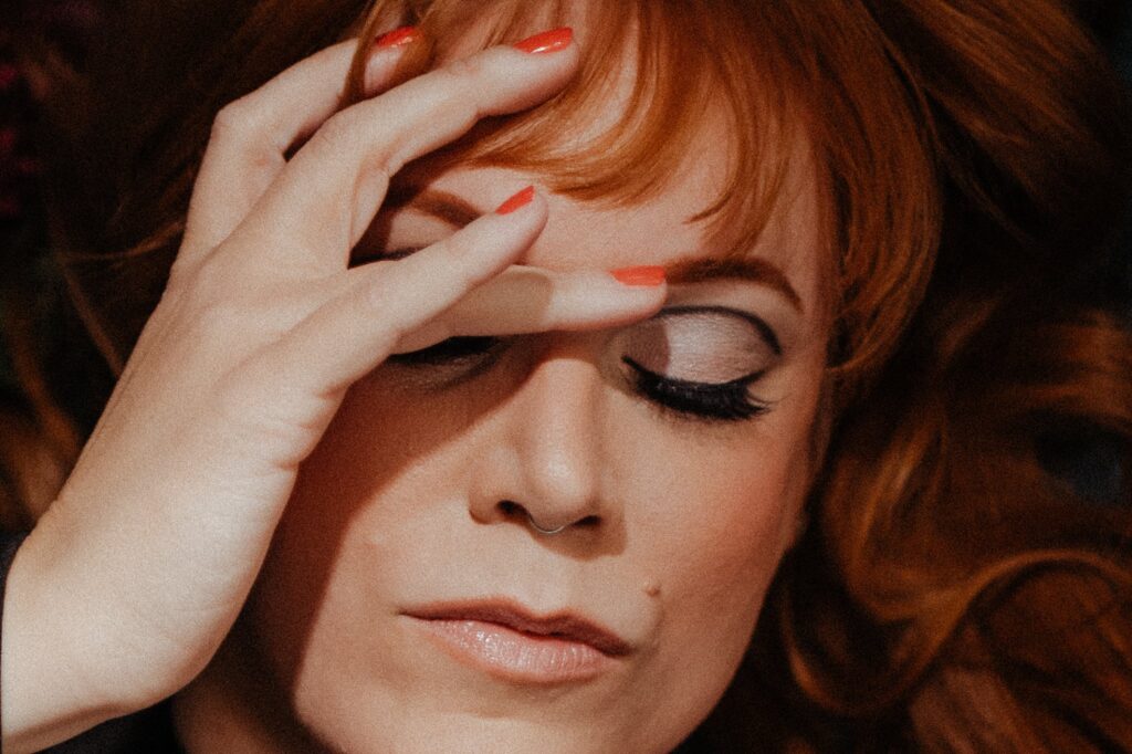 A person with red hair and coral nail polish touches their forehead, eyes closed, showcasing dramatic eye makeup and a serene expression—captured in the signature style of a Nashville creative portrait photographer.
