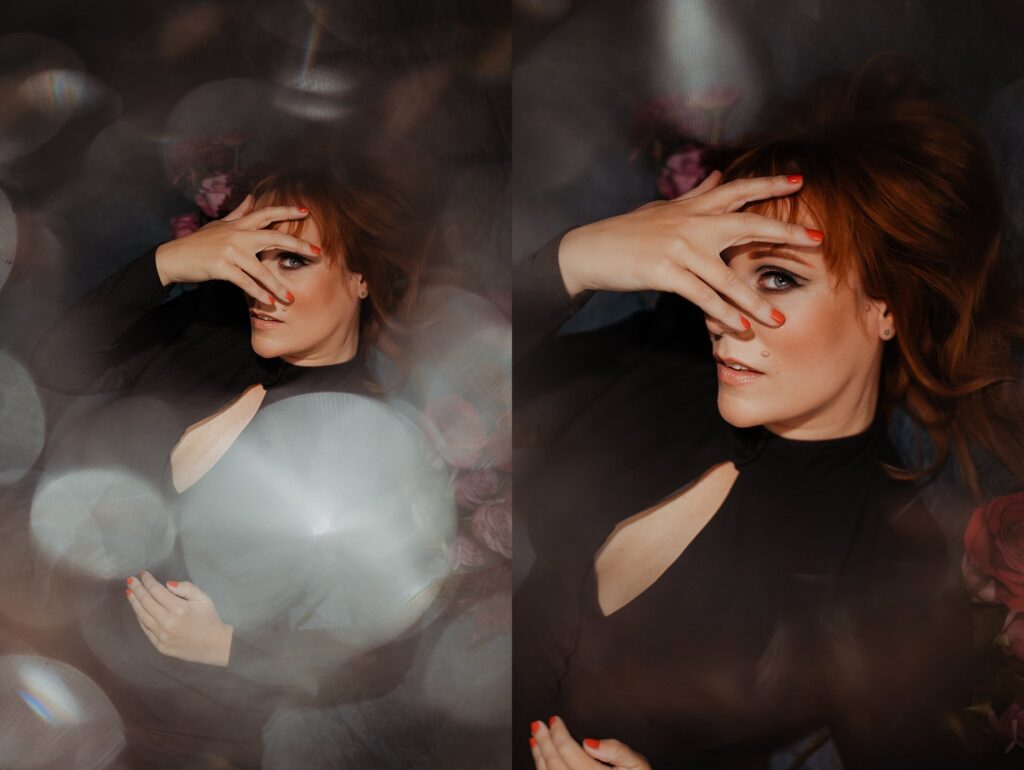 A woman with red hair lies on her back, wearing a black dress. She covers part of her face with her hand, showing red nails. Captured by a Nashville creative portrait photographer, the dreamy scene glows with blurred lights and roses around her.