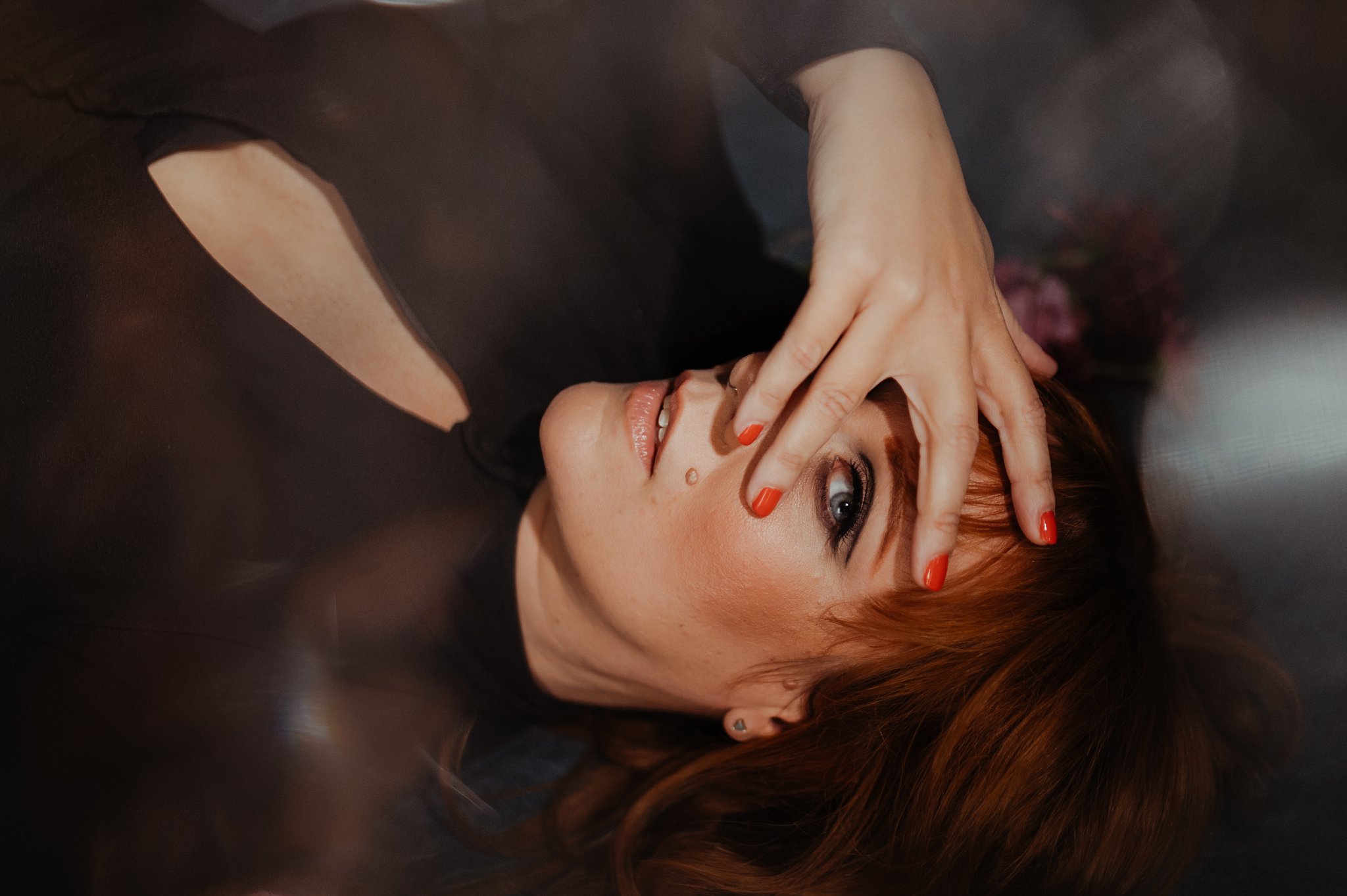 A woman with red hair and red nail polish lies on her back, gazing at the camera through her fingers, her hand partially covering her face. Captured by a Nashville creative portrait photographer, soft light creates a dreamy atmosphere.