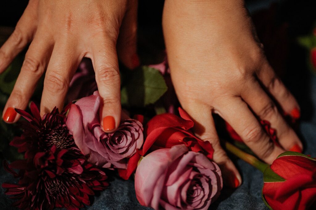 Two hands with red-painted nails rest gently on a mix of red and purple flowers, including roses and chrysanthemums, against a dark background—a striking image by a Nashville creative portrait photographer.