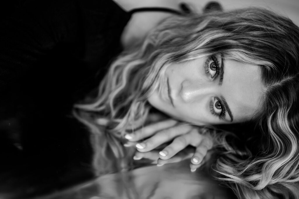 A black-and-white close-up by a Nashville creative portrait photographer captures a woman lying down with her head on a reflective surface. Her wavy hair frames her face as she gazes intently at the camera, with her hand resting nearby.