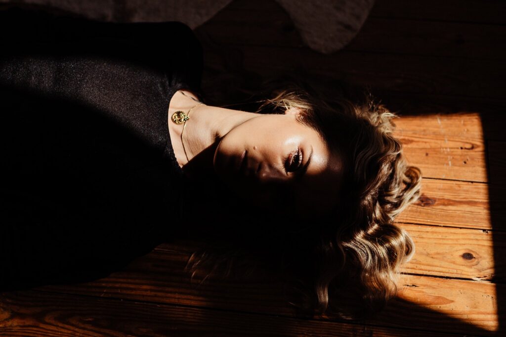 A woman with wavy blonde hair lies on a wooden floor, partly in shadow and sunlight. Captured by a Nashville creative portrait photographer, she wears a black top and gold necklace, the light perfectly highlighting one side of her face.