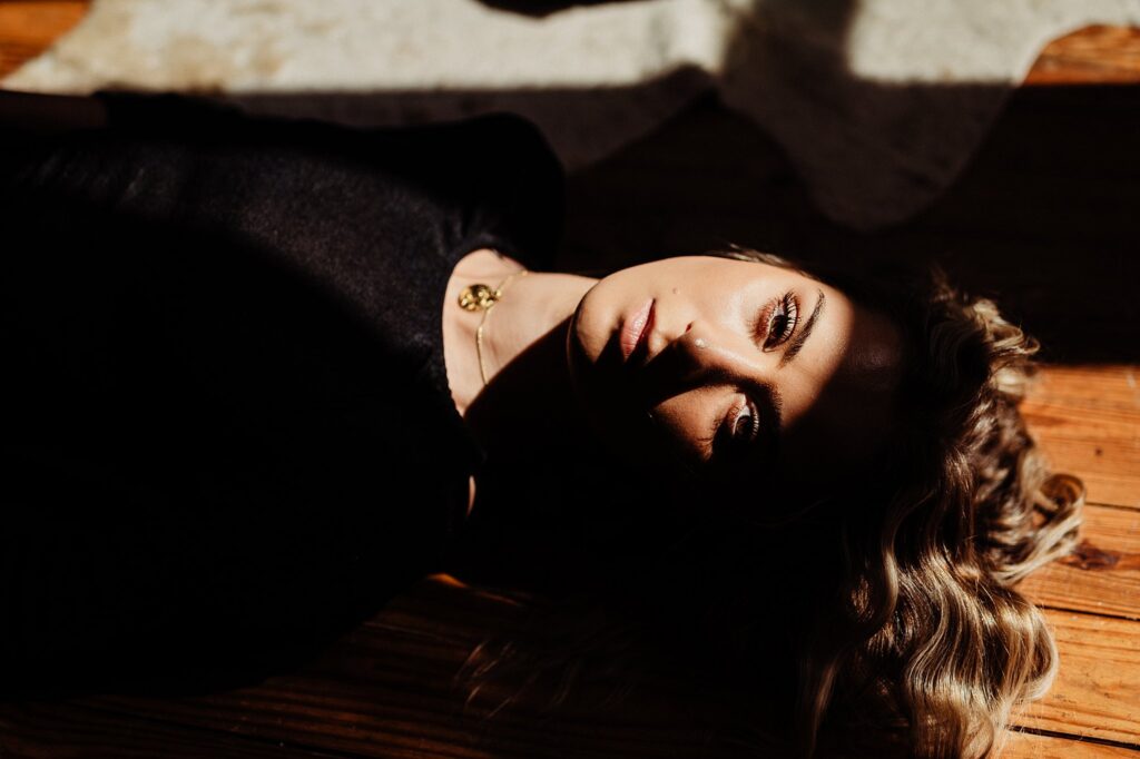 A woman with wavy blonde hair lies on a wooden floor, partially in shadow and sunlight. She wears a black top and a gold necklace, gazing softly at the camera—a striking image by a Nashville creative portrait photographer.