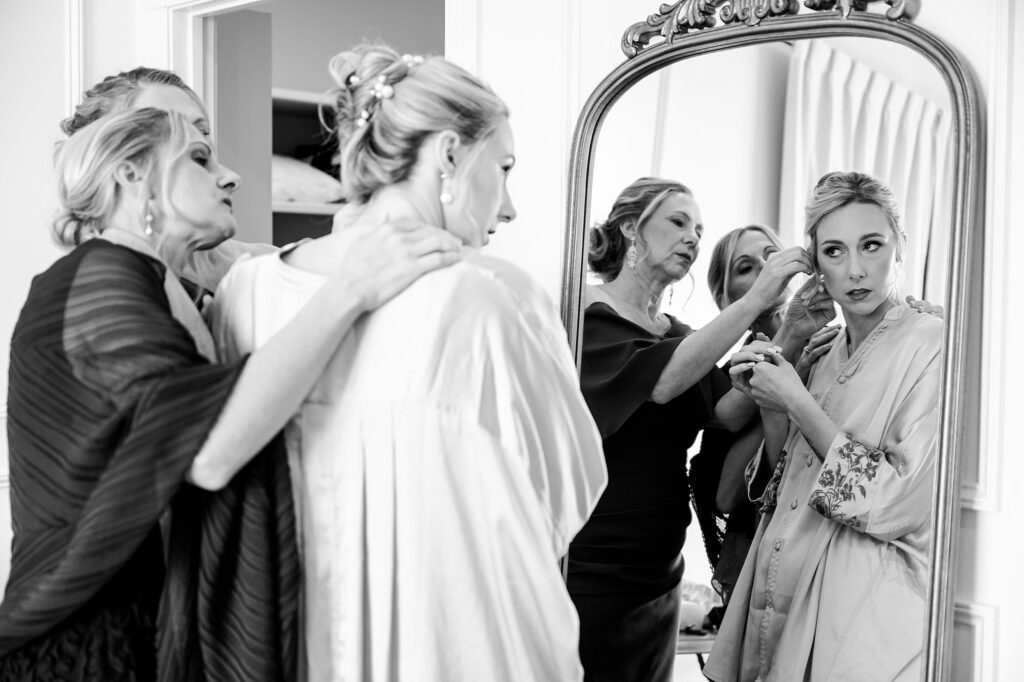 Three women help another woman adjust her hair and robe in front of a mirror. The intimate, caring scene—captured by a Nashville wedding photographer—shows them in elegant attire, suggesting special occasion preparations.