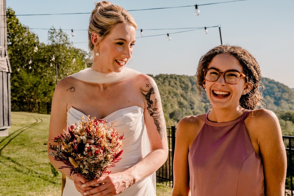 Two women smiling outdoors at a wedding; one in a white dress holding a bouquet, the other in a mauve dress with glasses. Captured by a Nashville wedding photographer, string lights and green hills glow under the clear sky.