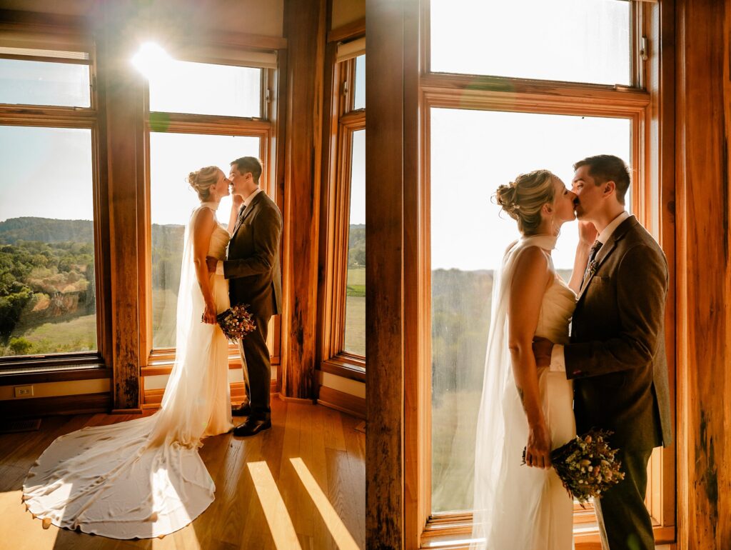 A bride and groom stand in front of large sunlit windows, sharing a kiss as warm sunlight streams into the wooden room. Captured by a Nashville wedding photographer, the moment highlights her long white dress and beautiful bouquet.