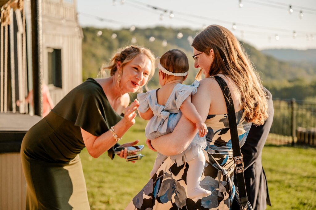 Two women stand outdoors in sunlight, one holding a baby in a blue dress with a white headband. The other woman smiles and waves at the baby. String lights and green hills set the scene—a perfect moment for a Nashville wedding photographer to capture.