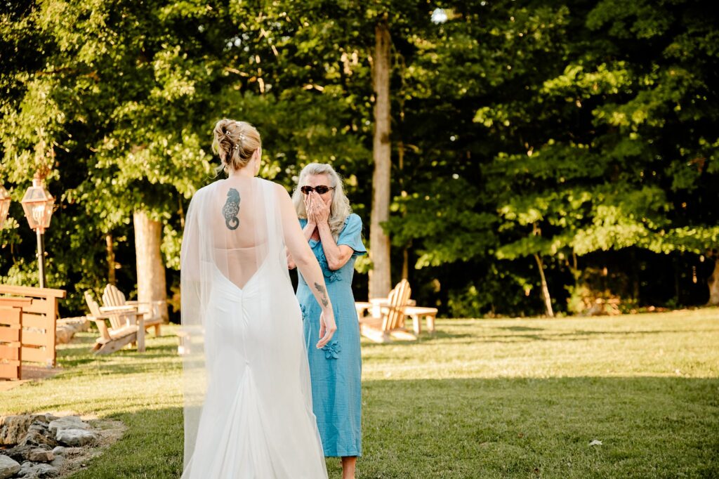 A bride in a white dress with a back tattoo stands on grass, facing an older woman in a blue dress who covers her mouth in surprise and joy—captured beautifully by a Nashville wedding photographer among trees and wooden chairs.
