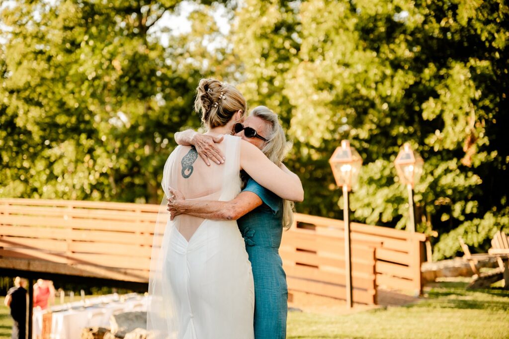 A bride in a white dress hugs an older woman in a blue dress and sunglasses outdoors, near a wooden bridge and trees, on a sunny day, beautifully captured by a Nashville wedding photographer.