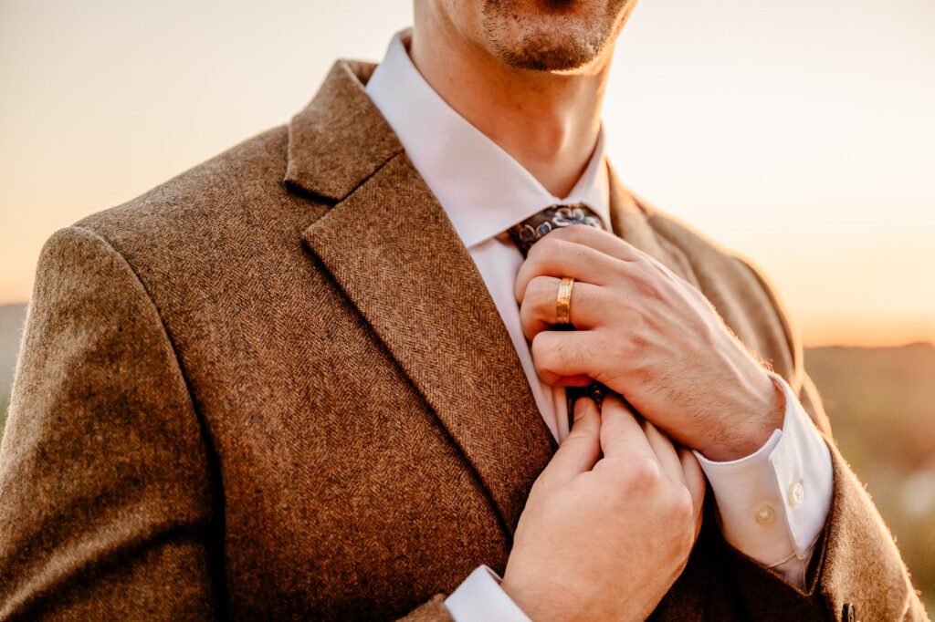 A man in a brown suit adjusts his patterned tie at sunset, wearing a white shirt and a gold wedding ring—captured perfectly by a Nashville wedding photographer.
