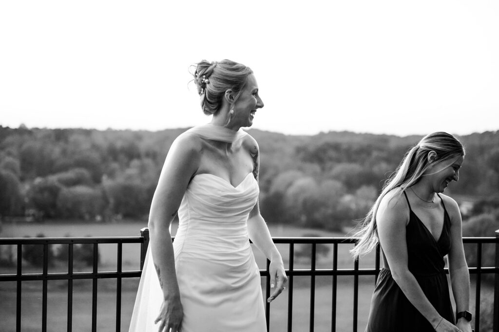 Two women stand on an outdoor balcony overlooking a blurred, tree-filled landscape. Captured by a Nashville wedding photographer, one smiles in a strapless wedding dress while the other, in a dark dress, laughs and looks down. The image is in black and white.