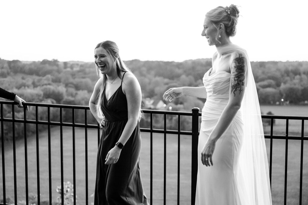 Two women stand laughing by a black metal railing overlooking a scenic landscape; one in a dark dress, the other in a white wedding gown with a sheer cape and visible arm tattoo. Captured in black and white by a Nashville wedding photographer.