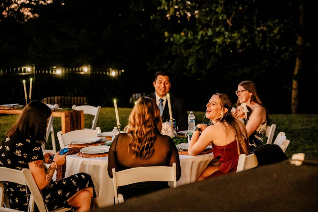 A group of five people sit around a round table outdoors at dusk, enjoying conversation and laughter. Captured by a Nashville wedding photographer, string lights glow in the background, creating a warm, festive atmosphere.