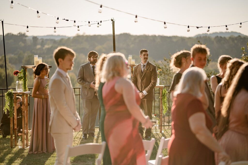 A group of people dressed formally gather outdoors at a wedding ceremony during sunset. String lights hang above, and trees and hills are visible in the background. Captured by a Nashville wedding photographer, some guests are seated while others stand.
