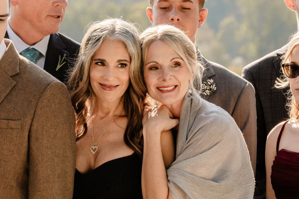 Two women smiling and standing close together, surrounded by others in formal attire at an outdoor event. Captured by a Nashville wedding photographer, one wears a heart necklace and black dress; the other, a light shawl with her hand on her friend's shoulder.