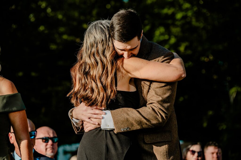 A man in a brown jacket and a woman in a black dress share a warm hug outdoors, surrounded by seated people under sunlight, as captured beautifully by a Nashville wedding photographer with trees in the background.