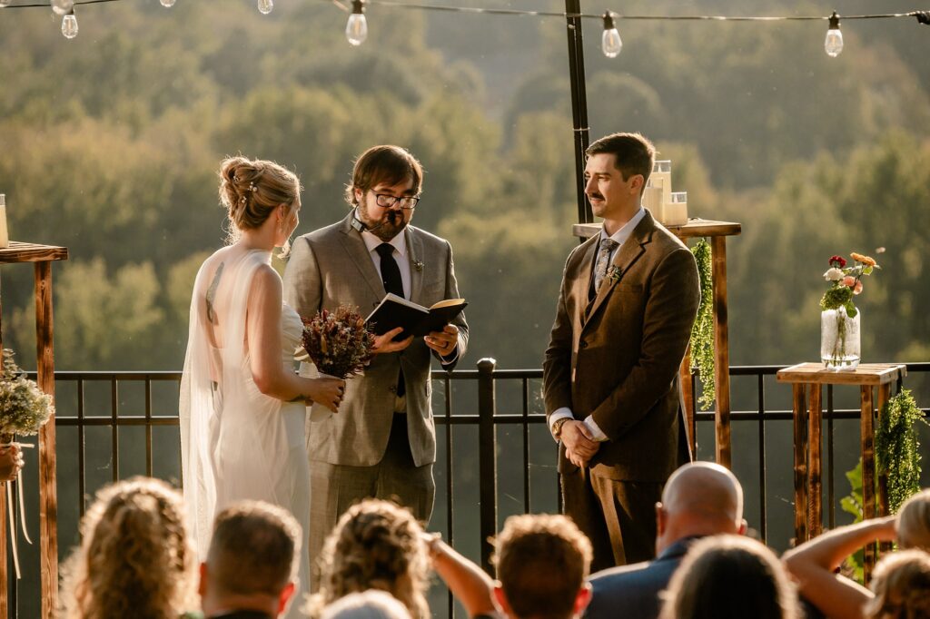 A bride and groom stand facing each other at an outdoor wedding ceremony, as a Nashville wedding photographer captures the moment. Guests watch as sunlight illuminates the scene, with trees and string lights in the background.