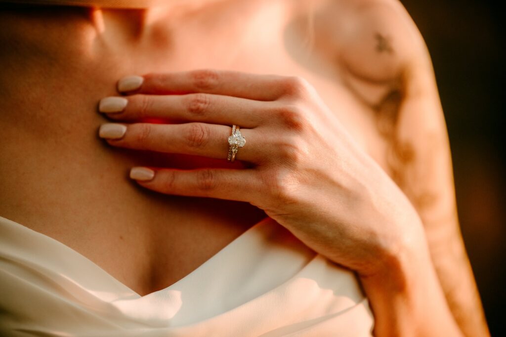 A close-up of a woman's hand resting on her chest, showing a diamond engagement ring. Captured by a Nashville wedding photographer, she has nude-colored nails and wears an off-the-shoulder white garment; part of a tattoo is visible on her upper arm.
