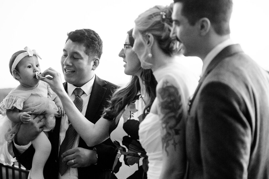 A man in a suit smiles as he feeds a baby girl with a bow headband, while three adults beside them, including a woman in a wedding dress, look on and smile. Captured in black and white by a Nashville wedding photographer.