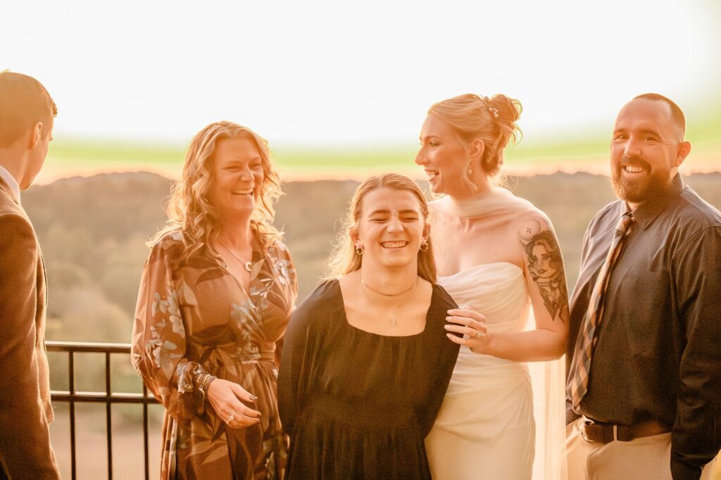 Five adults, dressed in formal attire, stand together outdoors at sunset, smiling and laughing. One woman wears a white dress, suggesting a wedding or celebration—captured beautifully by a Nashville wedding photographer against a blurred landscape and railing.