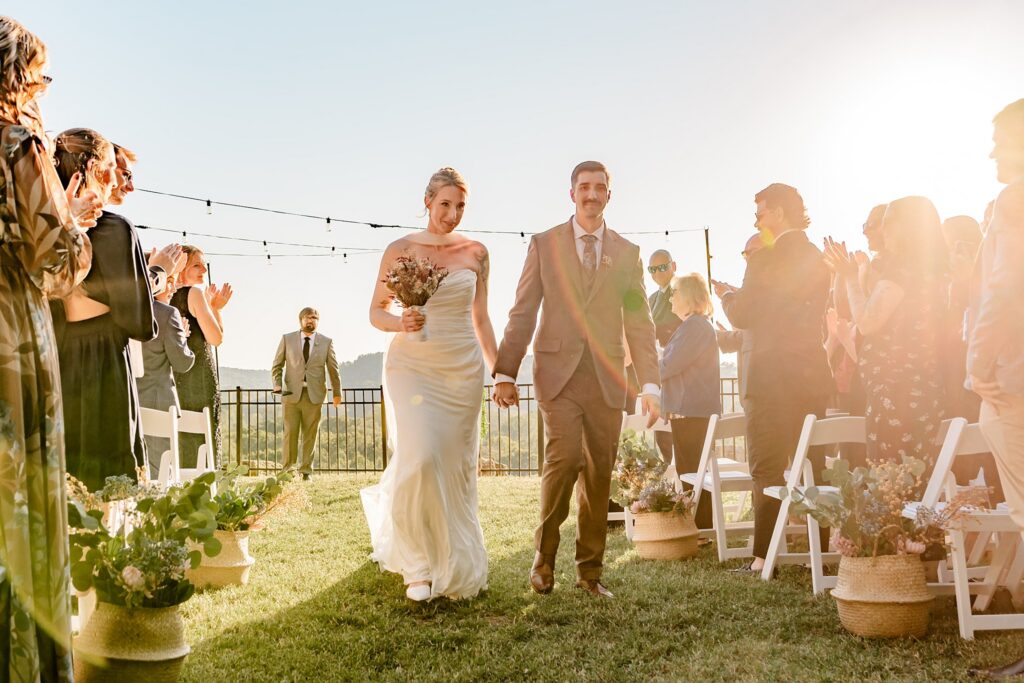 A bride and groom walk hand in hand down an outdoor aisle, smiling as guests on either side stand and applaud. Captured by a Nashville wedding photographer, the sun sets behind them, casting a warm glow over the celebratory scene.