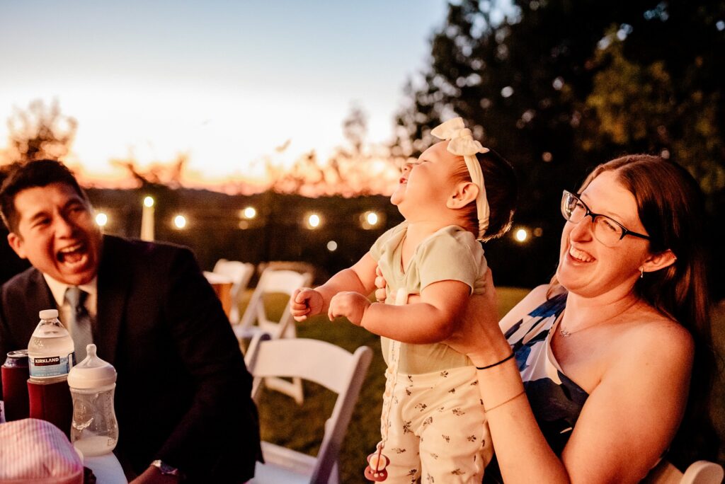 A woman holds a laughing baby in her lap while a man next to them smiles widely. Captured by a Nashville wedding photographer, they sit outdoors at sunset with string lights and trees in the background.