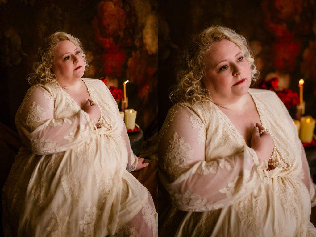 A person with curly blonde hair in a cream lace dress poses thoughtfully indoors, holding a pearl necklace. Captured by a Nashville creative portrait photographer, warm candlelight and deep red flowers create a soft, romantic background.