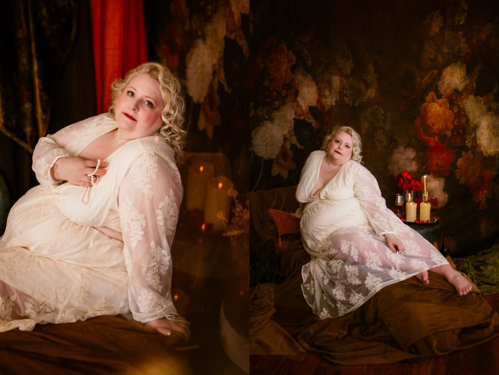 A woman in a sheer, white lace dress poses in a dim, romantic setting with floral wallpaper and red flowers. Captured by a Nashville creative portrait photographer, she holds a pearl necklace and gazes softly at the camera amidst draped fabrics and candles.