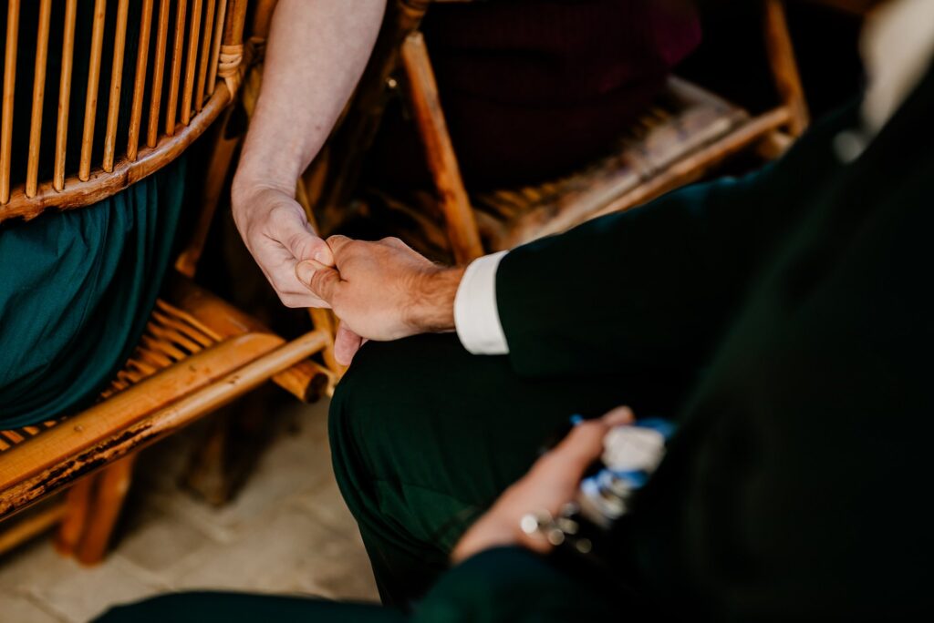 Two people sit side by side on wooden chairs, holding hands for comfort or support. One wears a dark suit, the other a light top. Captured with the artistry of a Nashville wedding photographer, the focus is on their clasped hands.