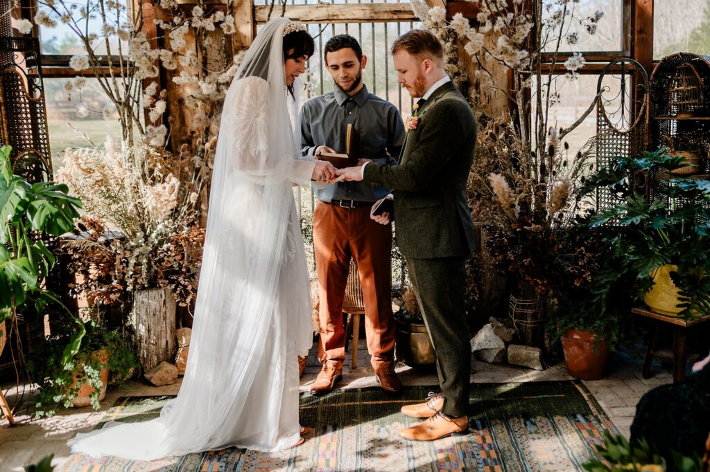 A bride and groom stand facing each other, holding hands, as an officiant reads vows inside a rustic, plant-filled room. Captured by a Nashville wedding photographer, light streams through large windows onto dried flowers and natural decor.