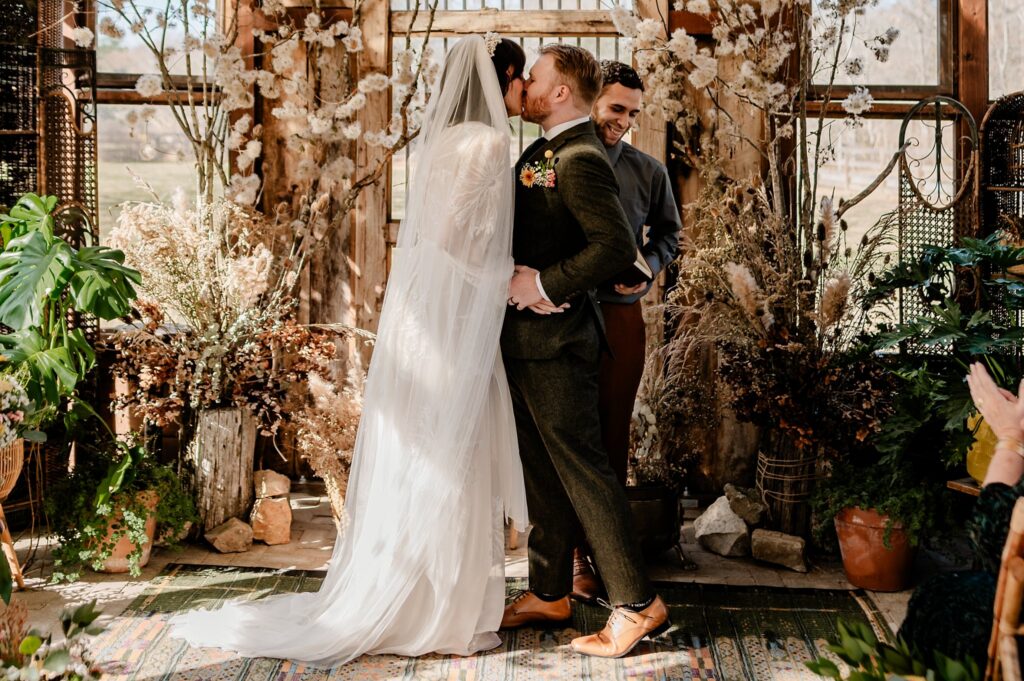 A bride and groom kiss at their wedding ceremony in a rustic, plant-filled venue, as their Nashville wedding photographer captures the moment, with an officiant smiling behind them and guests seated nearby.