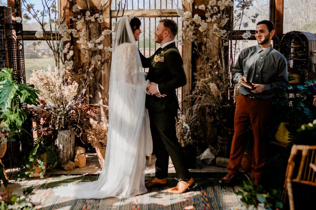 A bride and groom stand facing each other, smiling, during their wedding ceremony in a rustic, plant-filled setting as an officiant holds a book. Sunlight streams through large windows behind them, perfectly captured by a Nashville wedding photographer.