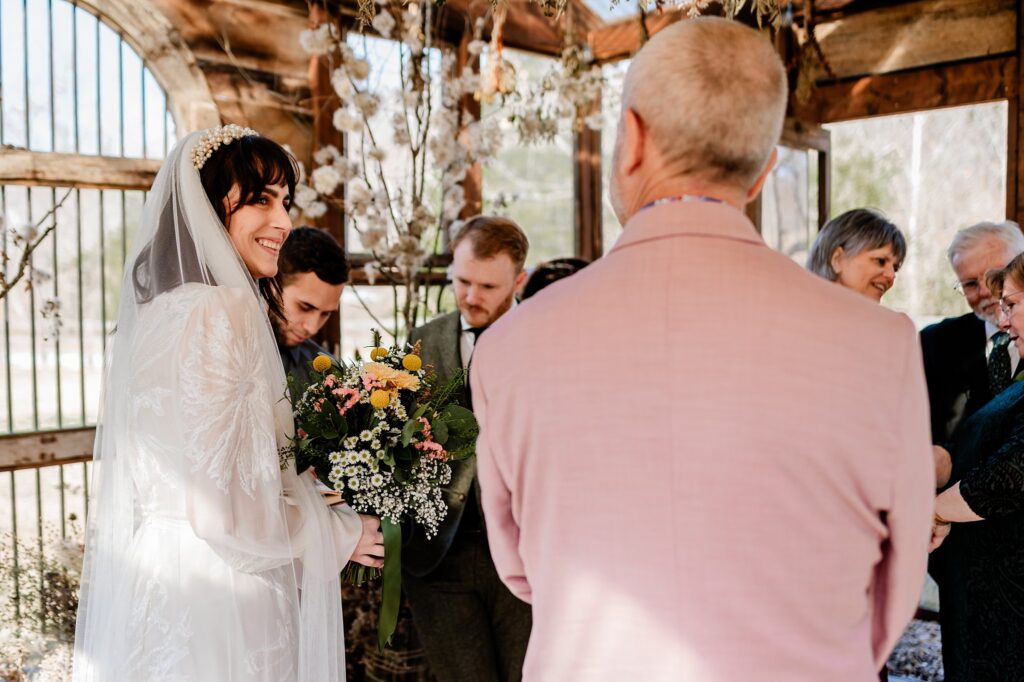 A smiling bride in a white dress and veil holds a bouquet of flowers during a wedding ceremony, beautifully captured by a Nashville wedding photographer in a rustic, sunlit setting with wooden beams and floral decorations.