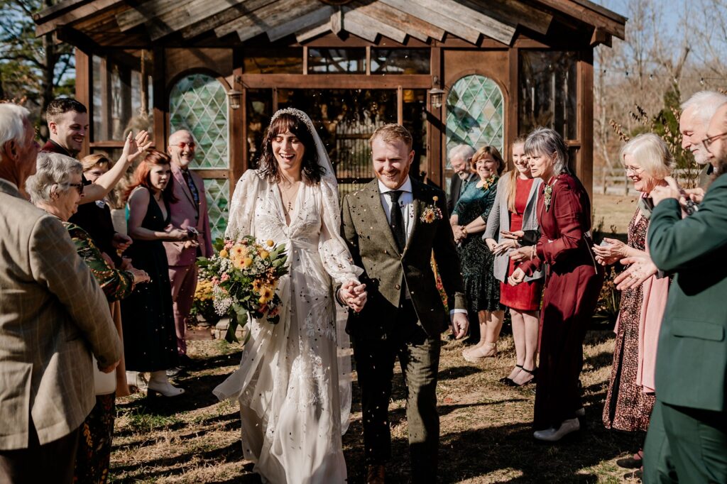 A bride and groom walk hand in hand outside a rustic wooden building, smiling as guests on both sides cheer and throw confetti. Captured by a Nashville wedding photographer, the joyful moment glows with love and laughter.