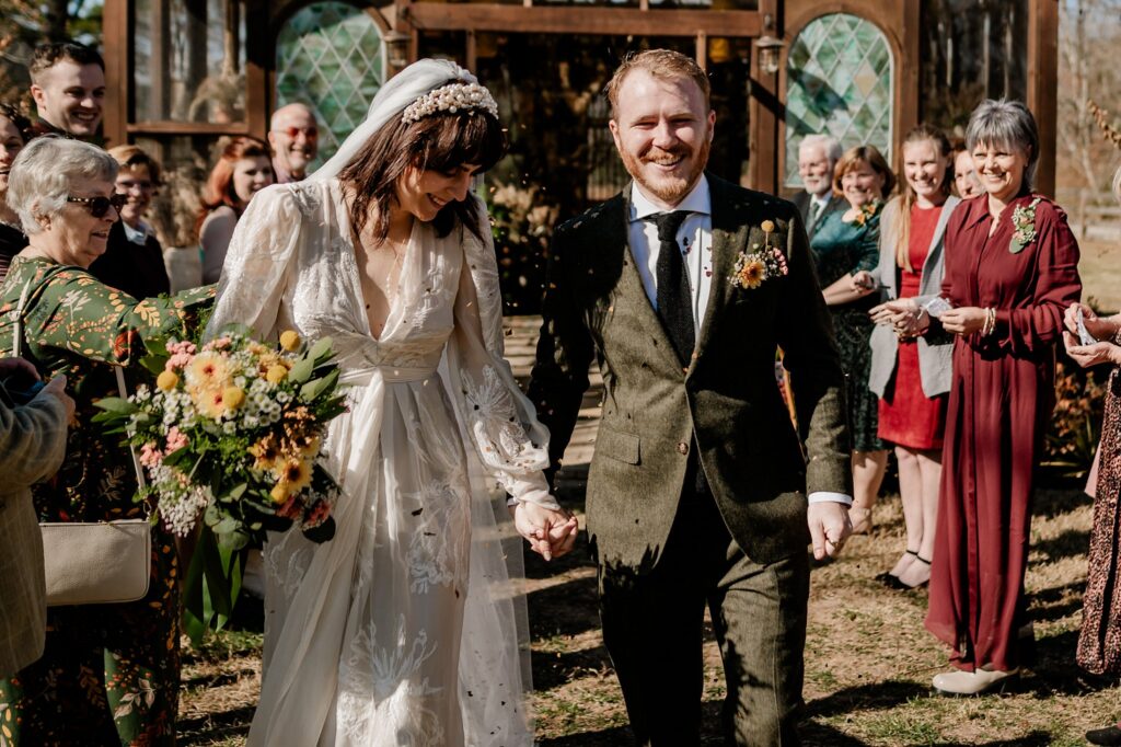 A smiling bride and groom walk hand in hand outside after their ceremony, surrounded by cheering guests. Captured by a Nashville wedding photographer, the sunlight highlights the bride’s bouquet and white dress as the groom beams in his green suit.