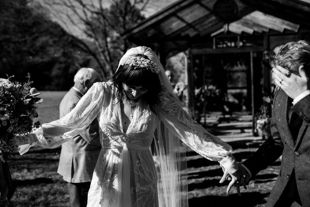 A bride in a long-sleeved lace gown and veil holds a bouquet, looking down while holding hands with her partner outside, as guests and a rustic building create a charming backdrop—captured by a Nashville wedding photographer.