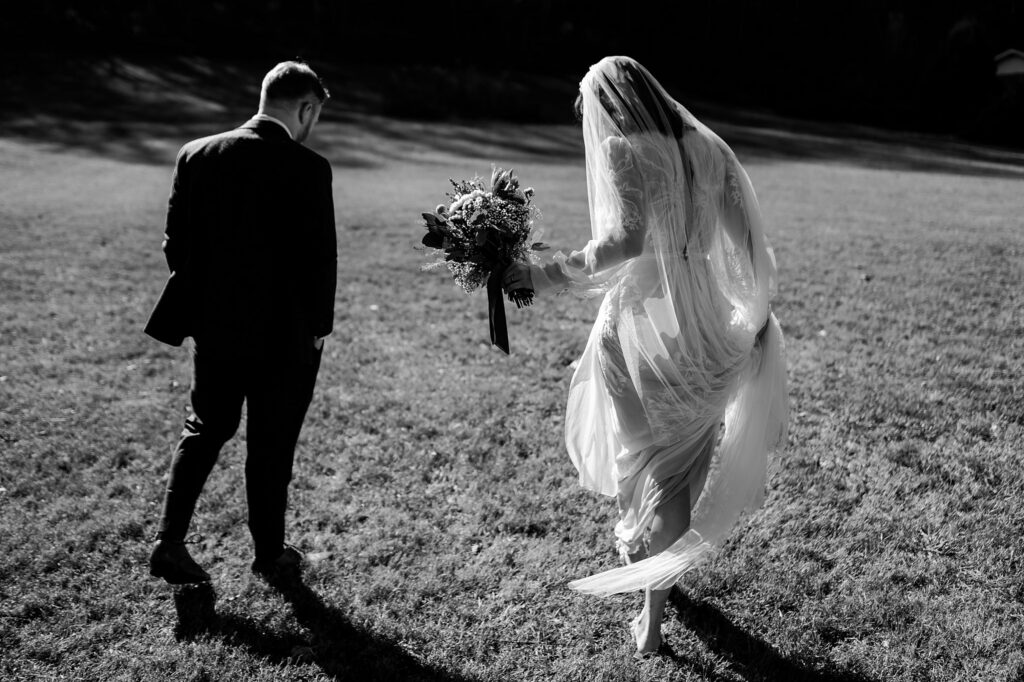 A bride in a flowing dress and veil walks barefoot on grass, holding a bouquet, beside a man in a suit. Captured in bright sunlight from behind by a Nashville wedding photographer.