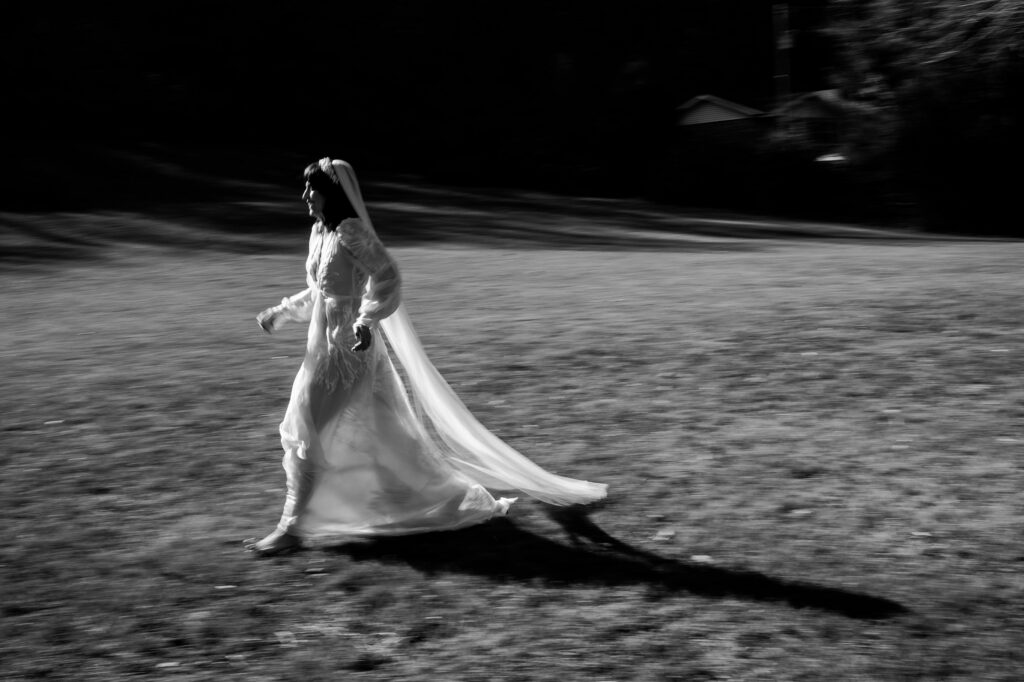 A person in a flowing white dress and veil walks quickly across a grassy field, casting a long shadow in the sunlight. Captured by a Nashville wedding photographer, the black-and-white photo is slightly blurred, emphasizing motion.