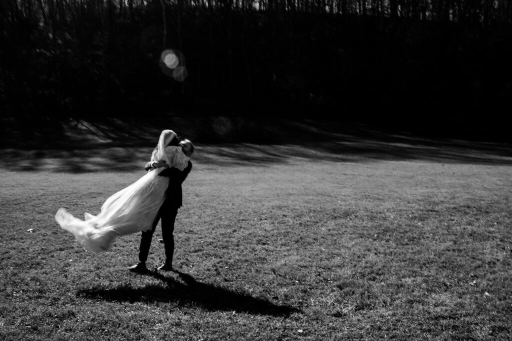 A person in a suit lifts and spins a person in a flowing dress on a sunlit grassy field, casting long shadows with trees behind them—a timeless black and white image by a Nashville wedding photographer.