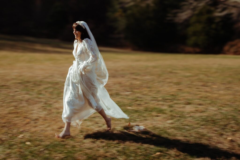 A barefoot bride in a flowing white dress and veil joyfully runs across a sunlit, grassy field, her dress trailing behind her—captured beautifully by a Nashville wedding photographer. The blurred background emphasizes her movement and happiness.