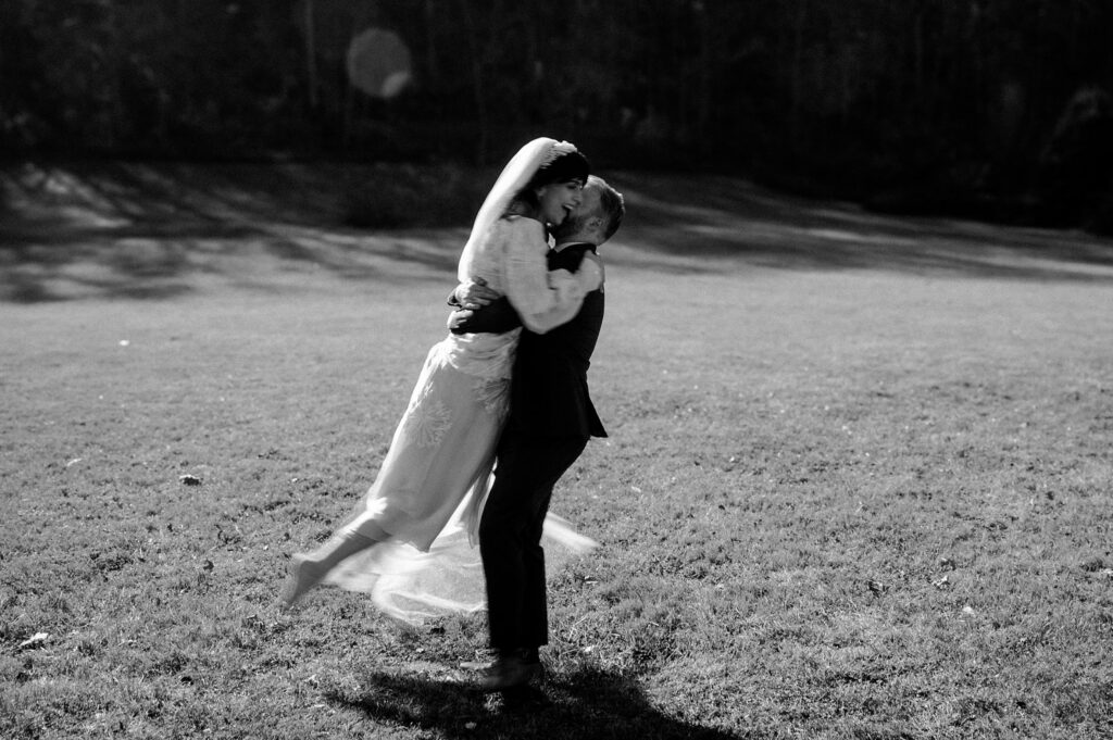 A person in a wedding dress is lifted and embraced by a person in a suit on a grassy field, both smiling joyfully. Captured in black and white, this moment reflects the artistry of a Nashville wedding photographer.