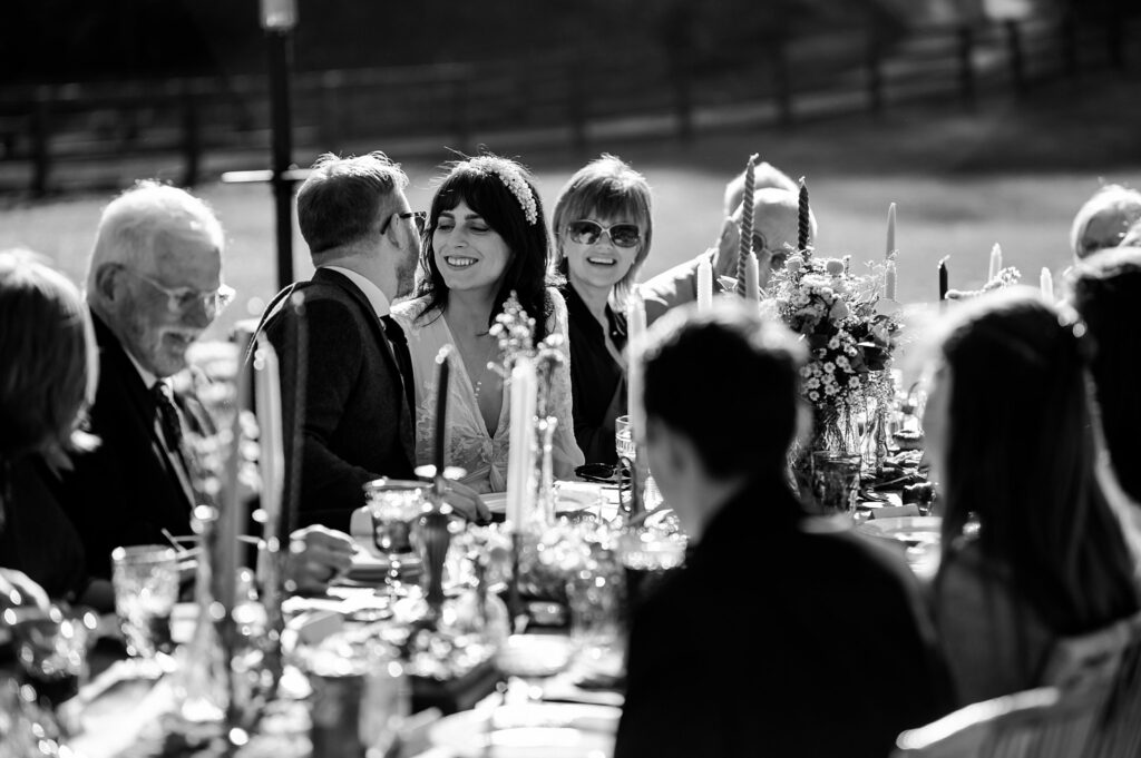 A group of people sit around a long outdoor table set for a meal, engaged in conversation and smiling. Captured by a Nashville wedding photographer, the black and white scene with natural light and a blurred background suggests a festive gathering.