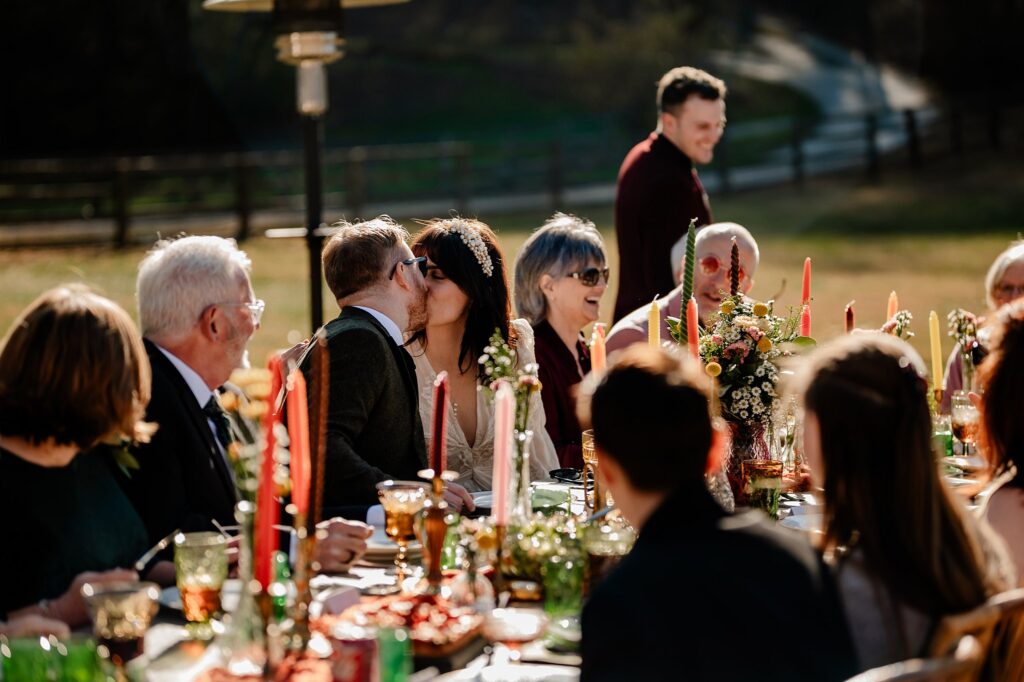 A bride and groom kiss at an outdoor wedding reception table, surrounded by guests enjoying food and drinks. Sunlight highlights the joyful scene as a Nashville wedding photographer captures colorful candles and flowers decorating the table.